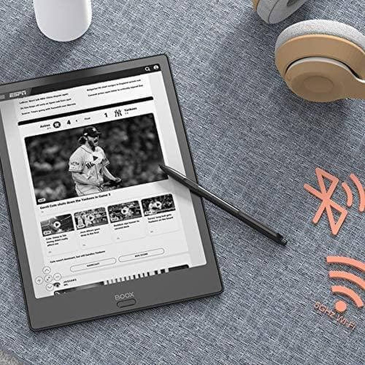 Tableta mostrando un partido de béisbol, con un lápiz óptico, auriculares y símbolos wifi/Bluetooth sobre superficie gris.