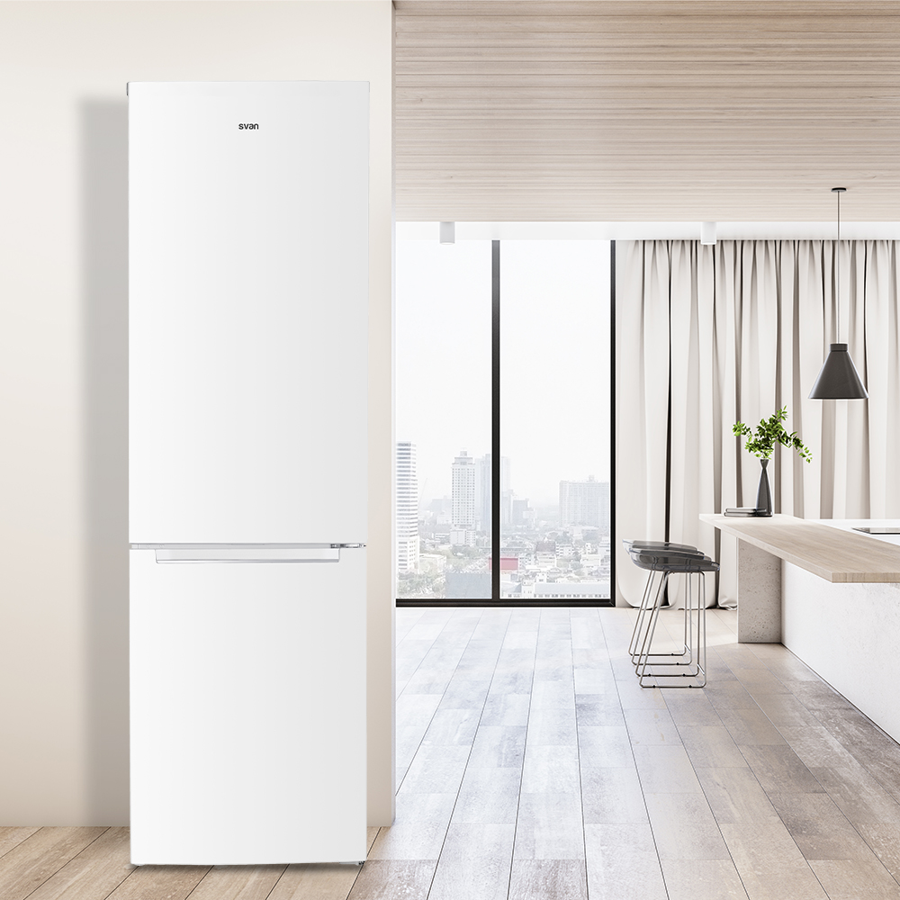 Un refrigerador blanco junto a una pared. En el fondo, una cocina con vista a la ciudad a través de una ventana grande.