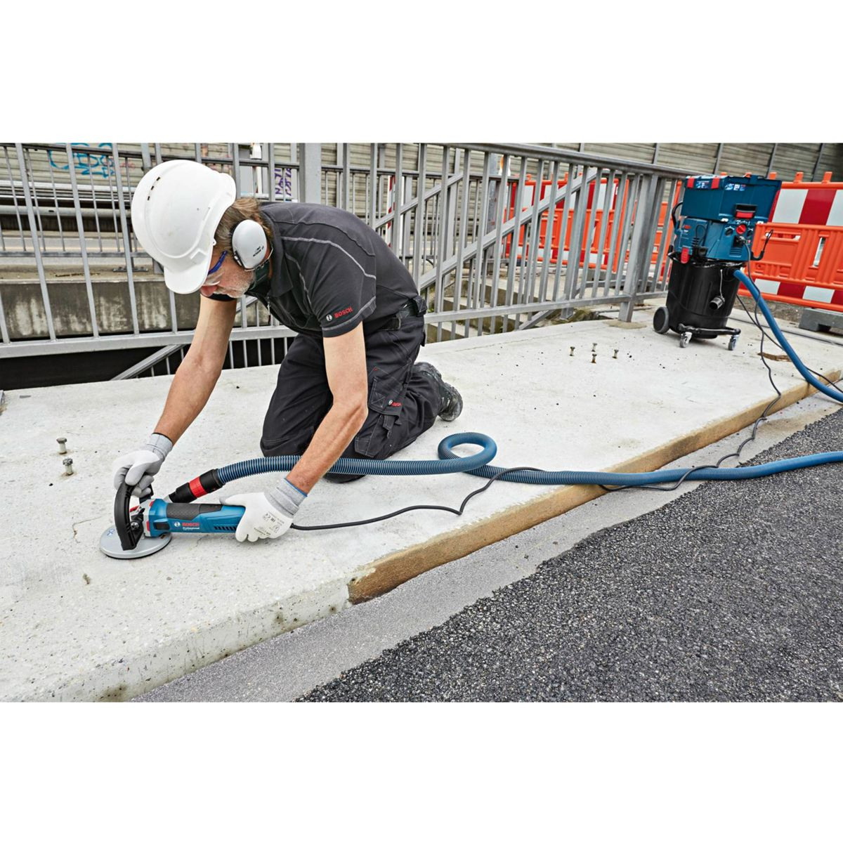 Ein Arbeiter kniet und schleift Beton mit einem Elektrowerkzeug, trägt Schutzkleidung. Ein Staubsauger und eine Barriere sind im Hintergrund.