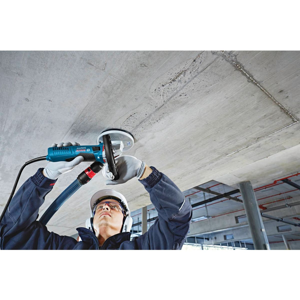 Ein Arbeiter verwendet ein Elektrowerkzeug an einer Betondecke. Er trägt Handschuhe, Helm und Gehörschutz. Das Werkzeug ist blau und schwarz.