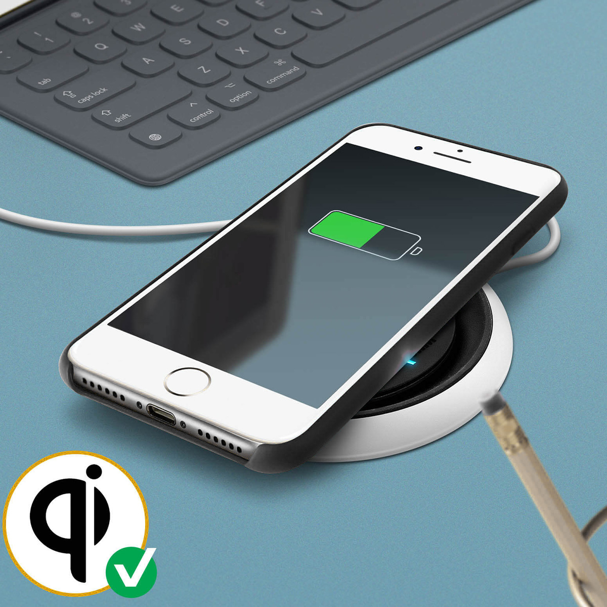 Uno smartphone bianco con custodia nera si ricarica in modalità wireless su un pad di ricarica bianco. Una tastiera sullo sfondo.