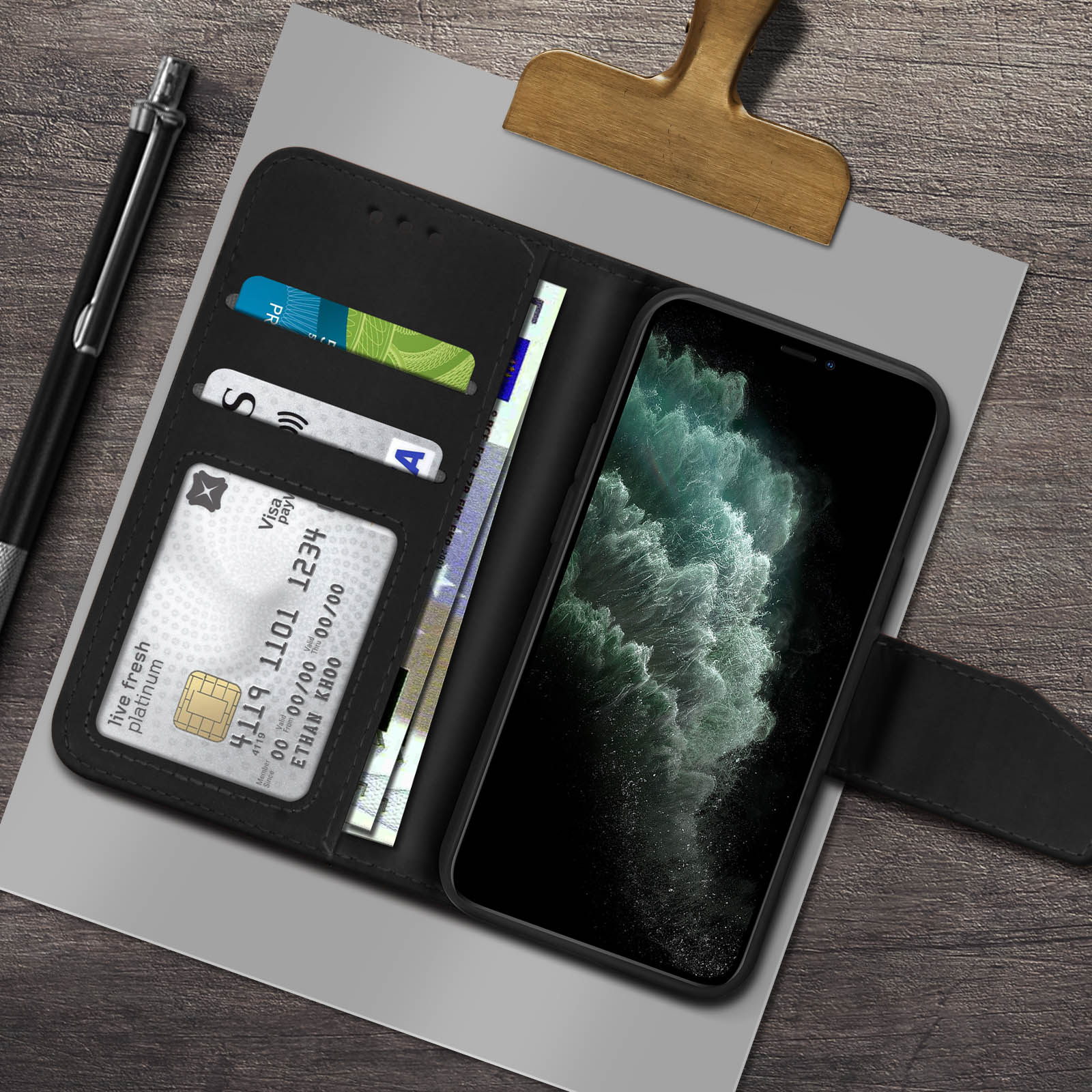 Una custodia nera per telefono con telefono, portafoglio con carte e denaro e una penna su una superficie di legno.