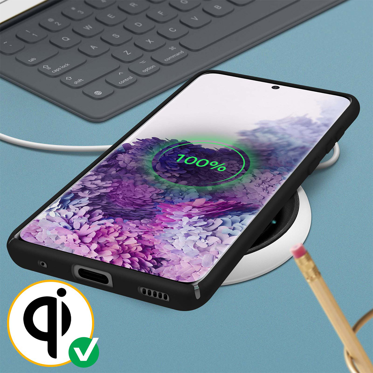 Smartphone in carica wireless, mostra il 100%. Tastiera e matita sullo sfondo. Logo Qi presente.