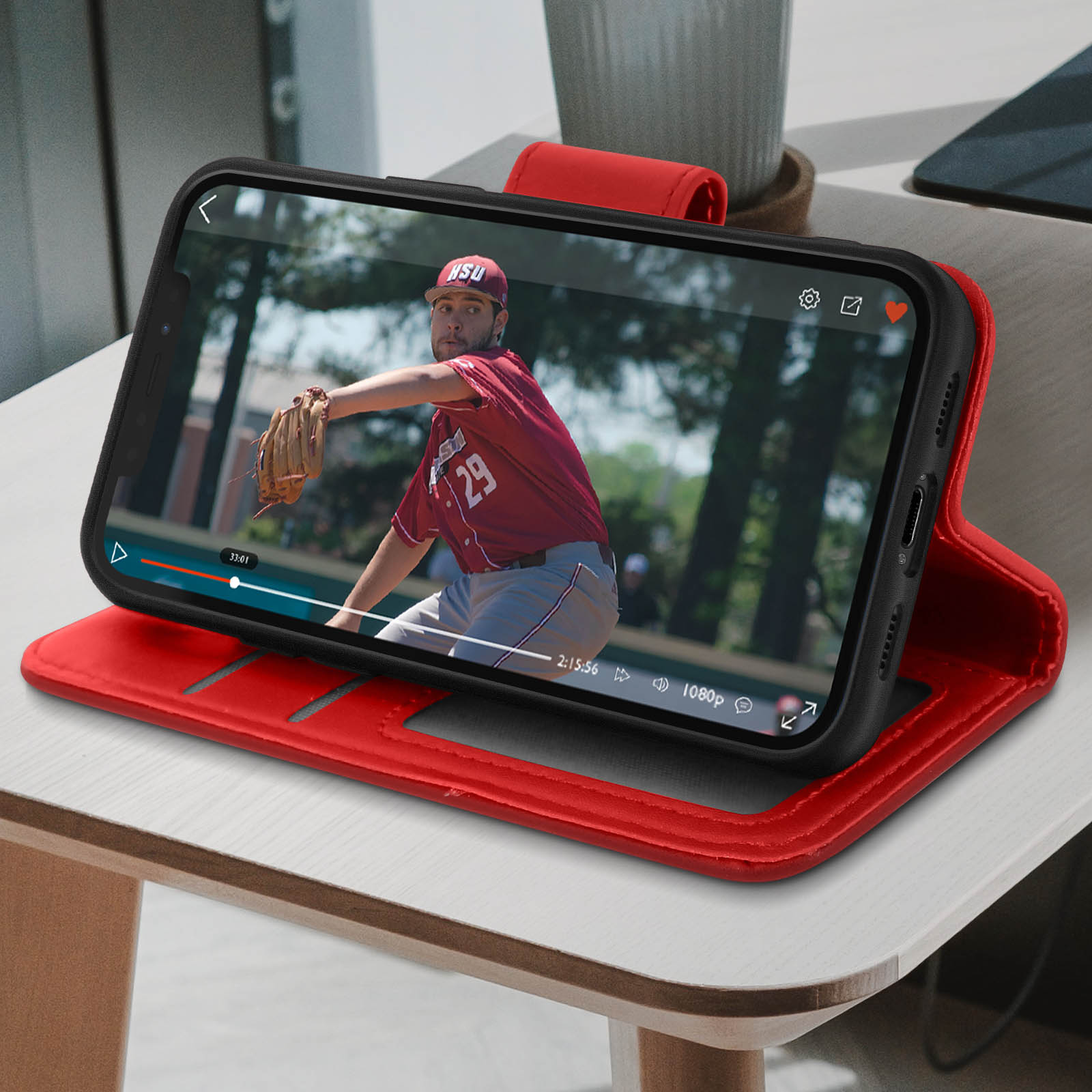 Telefono che mostra video di baseball in custodia rossa. Giocatore in uniforme bordeaux che lancia. Custodia su tavolo di legno.