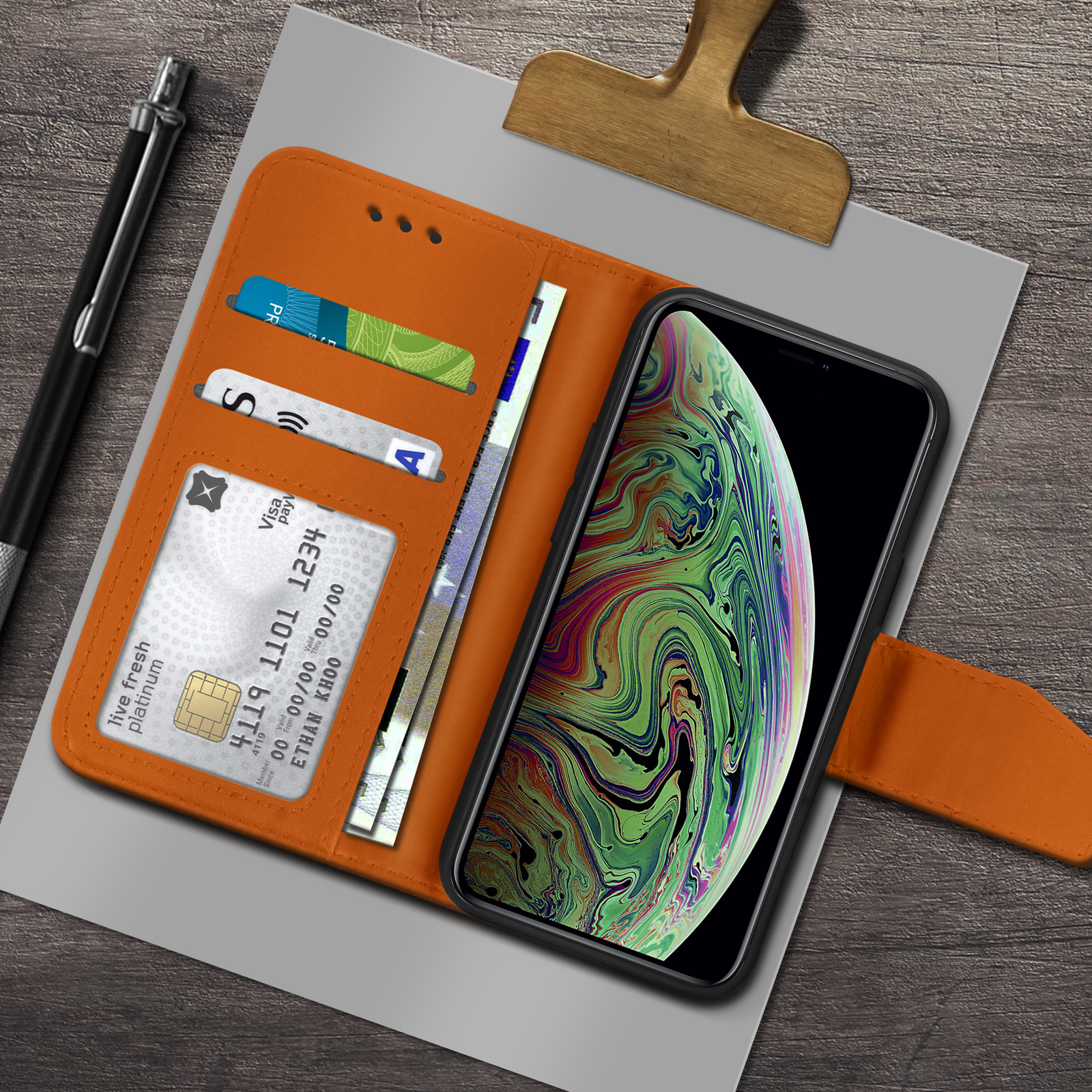 Un portafoglio arancione contiene carte e denaro accanto a un telefono con schermo colorato, su una superficie di legno con una penna.