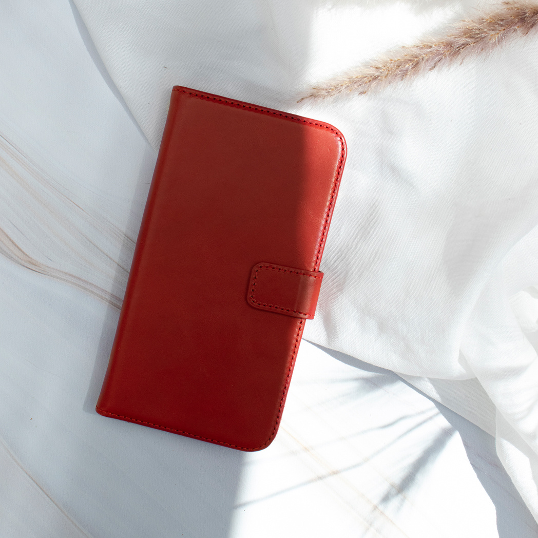 Rode leren telefoonhoes op witte stof met een stengel van gedroogd gras. De hoes heeft een magnetische sluiting.