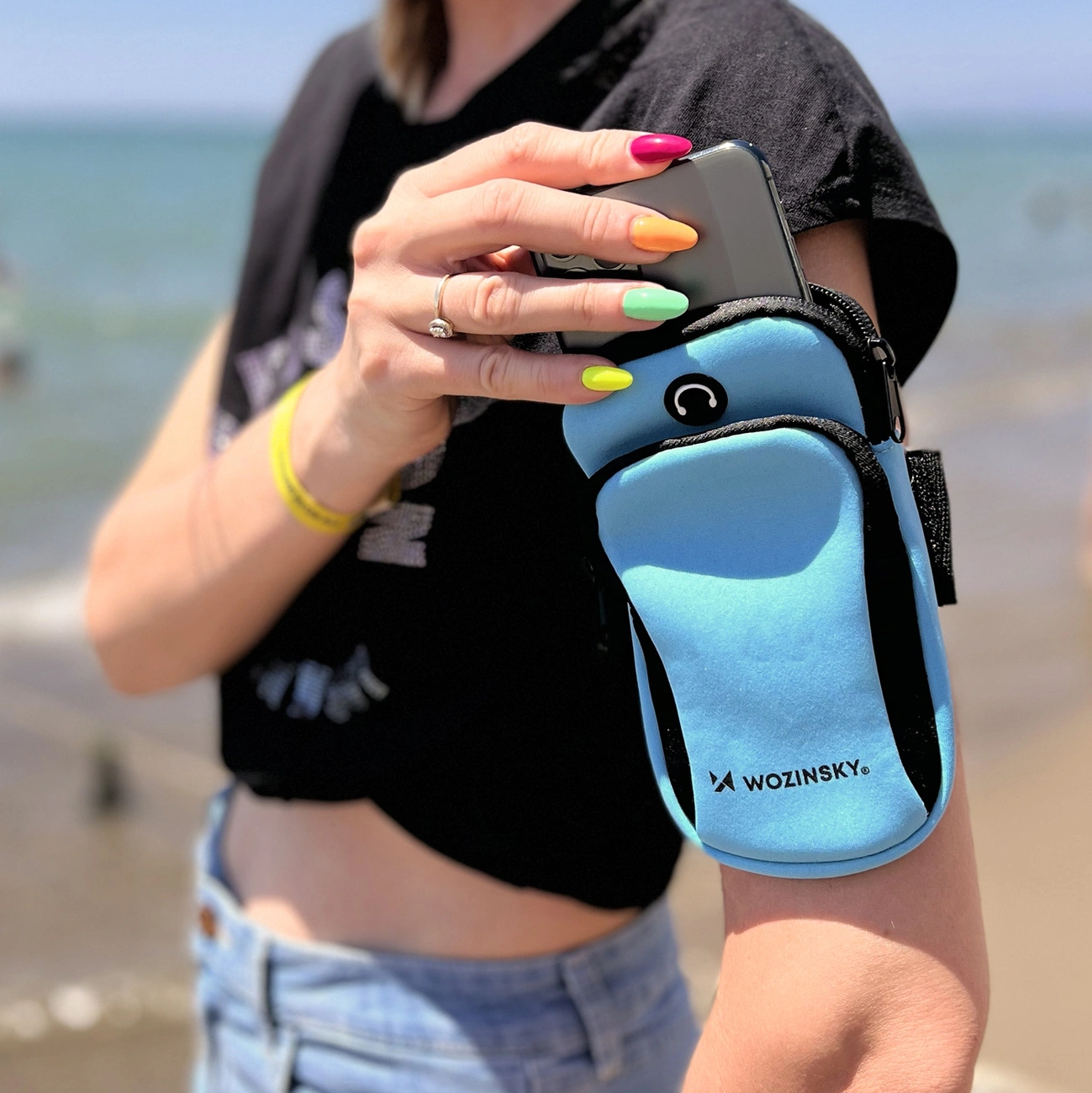 Eine Frau hält ein Telefon in einem blauen Armband. Ihre Nägel sind bunt. Der Hintergrund zeigt einen Strand und ein Meer.