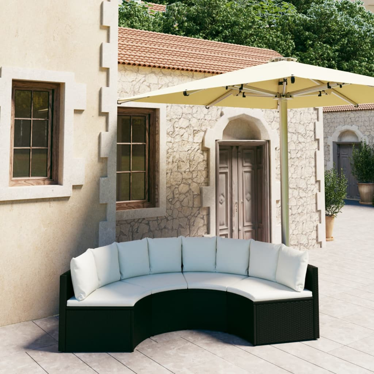Ein geschwungenes, schwarzes Outdoor-Sofa mit weißen Kissen steht unter einem beigen Schirm. Steinbauten und Bäume sind im Hintergrund.