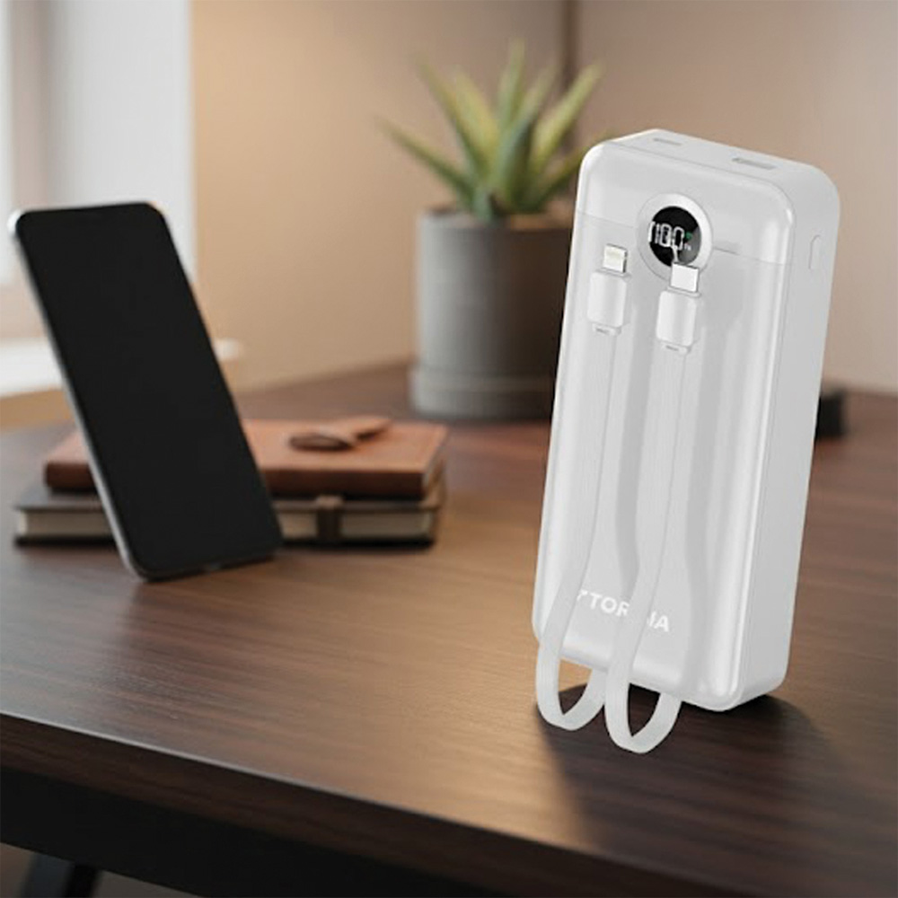 Ahşap bir masa üzerinde iki kablo bağlı beyaz bir powerbank ve bir telefon. Arka planda bir bitki ve bir kitap var.
