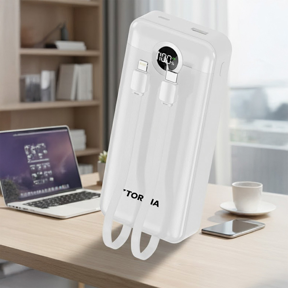 Beyaz powerbank, kablolar, %100 şarj, ahşap masa üzerinde. Arka planda dizüstü bilgisayar, telefon ve fincan da var.