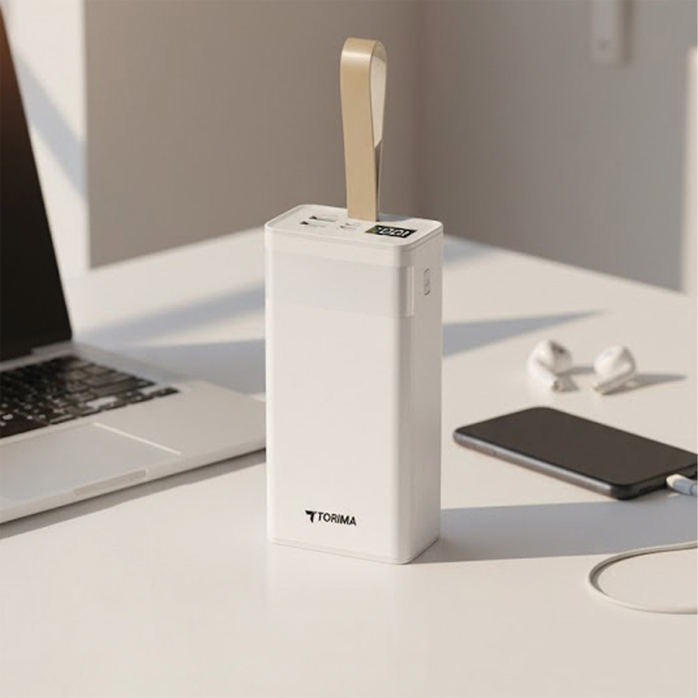 Bej renkli bir tutamağa sahip beyaz bir powerbank beyaz bir masada duruyor. Dizüstü bilgisayar, telefon ve kulaklıklar da var.