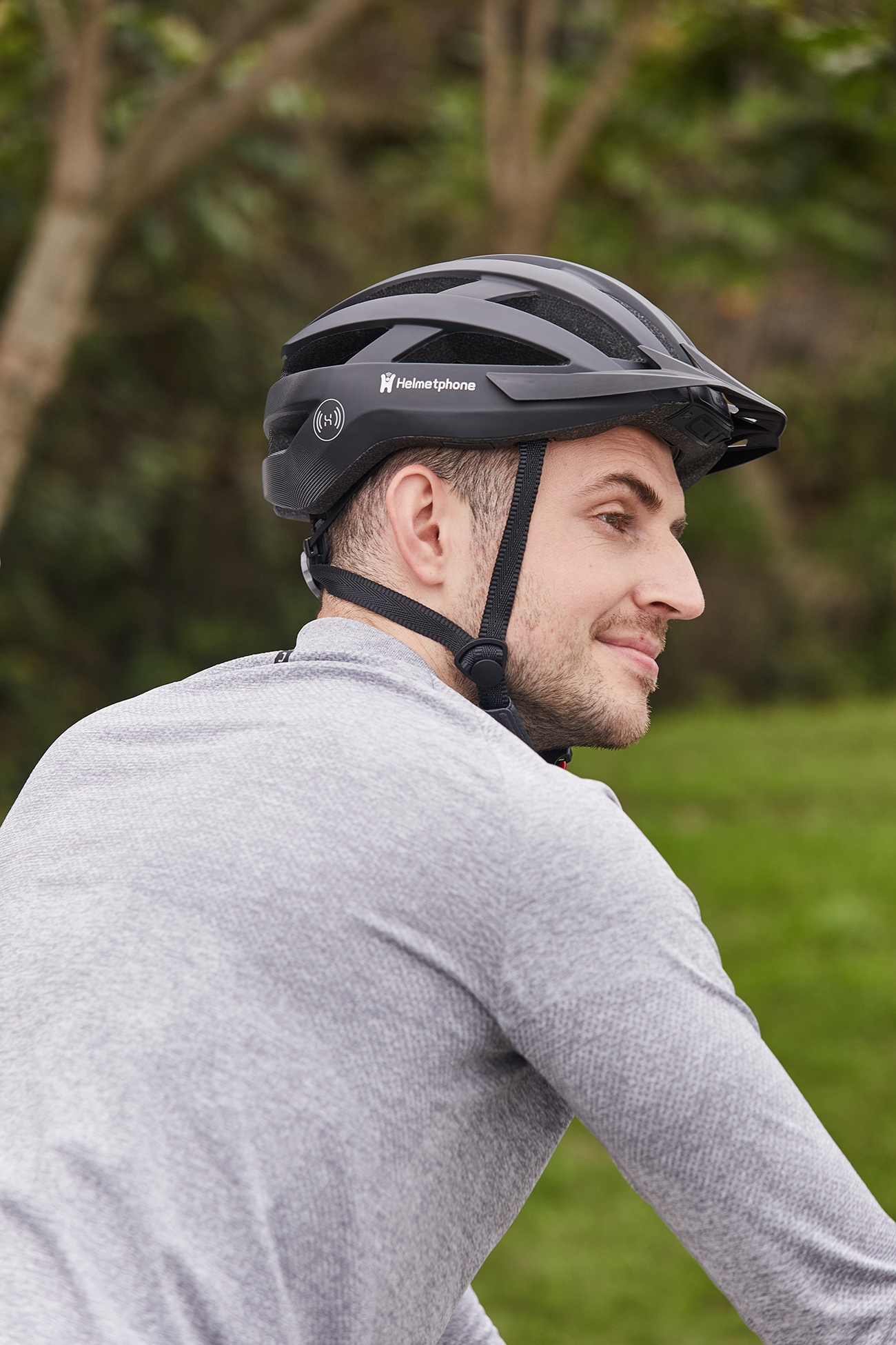 Uomo con casco nero con logo 'Helmetphone'. Indossa una camicia grigia, sorride, sfondo verde.