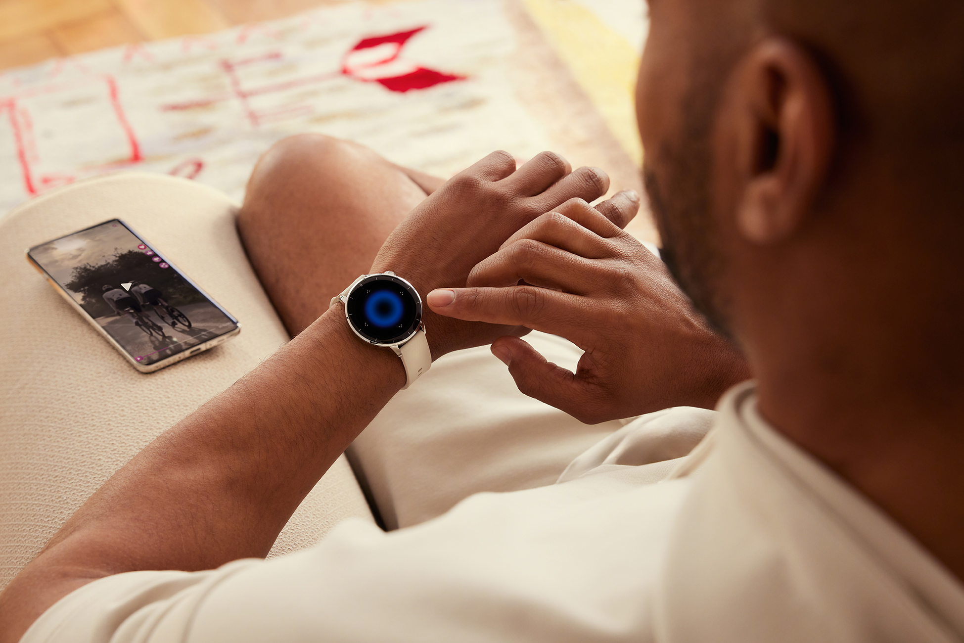 Mężczyzna patrzy na smartwatch. Telefon z kolarzami na ekranie leży obok zegarka. W tle widać dywan.