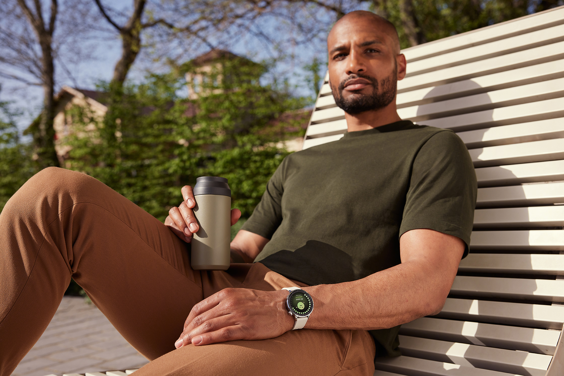 Łysy mężczyzna siedzi na ławce, trzymając kubek i nosząc smartwatch. Ma na sobie zieloną koszulę i brązowe spodnie, z drzewami w tle.