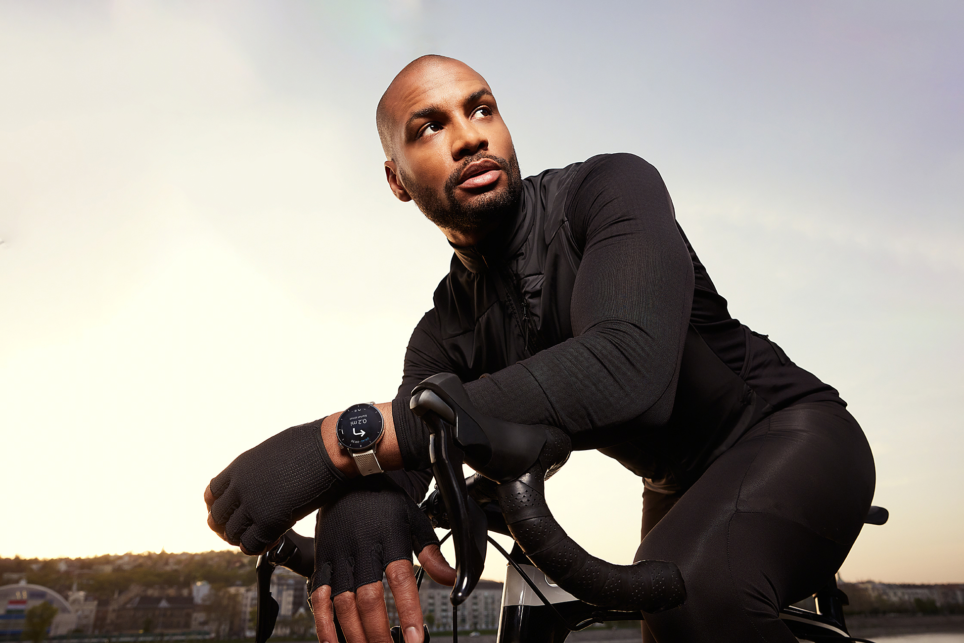 Mężczyzna w czarnym stroju sportowym opiera się o rower. Patrzy w górę i ma na sobie smartwatch. Tło: krajobraz miejski.