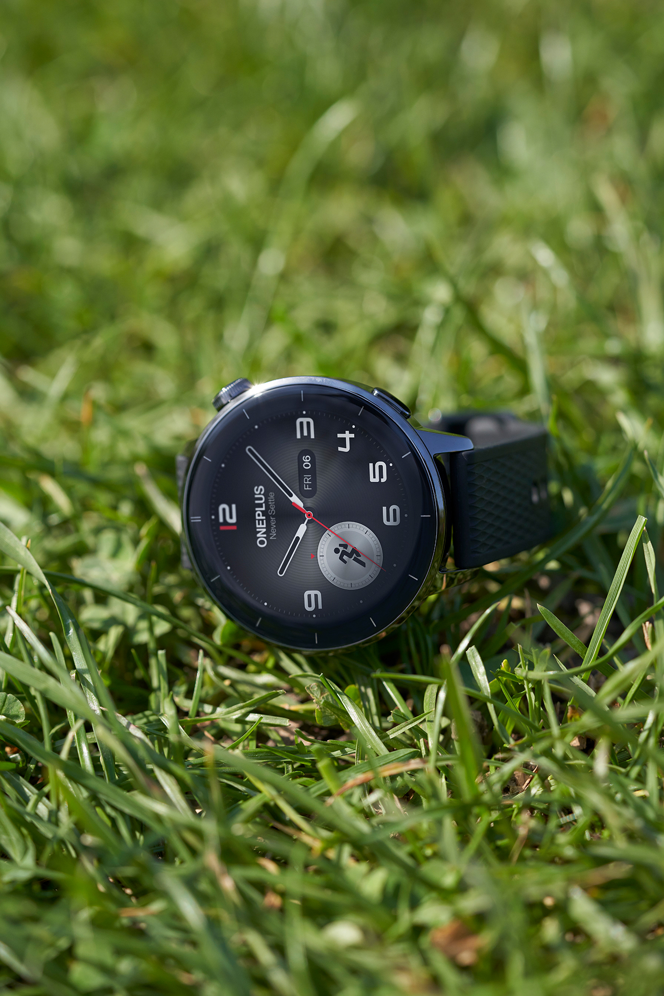 Un reloj inteligente OnePlus está sobre hierba verde. La esfera es negra con números y manecillas blancas. La correa es negra.