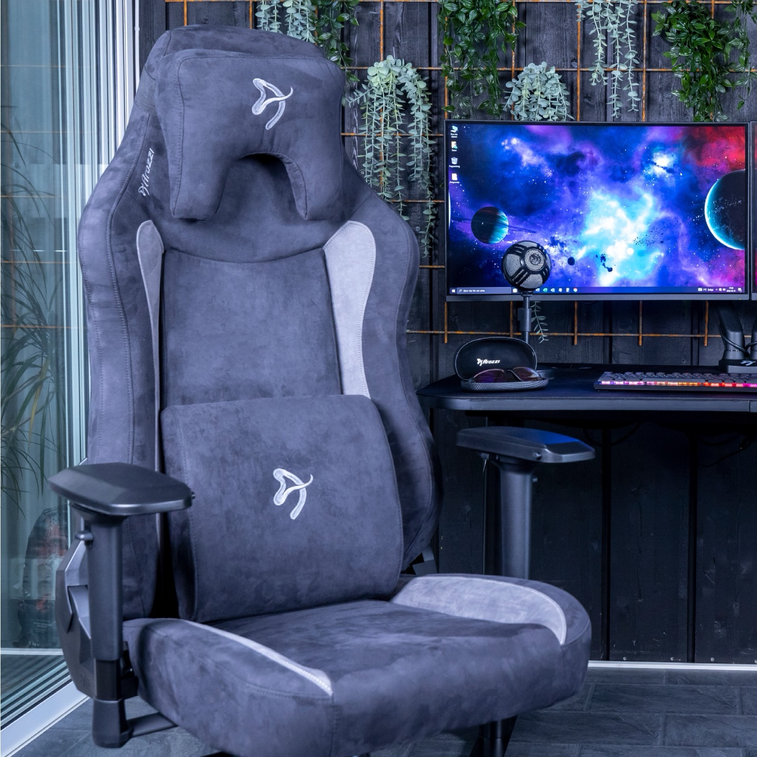 Een donkergrijze gamingstoel met hoofdsteun en lendenkussen. Een computermonitor toont een ruimtescène. Planten hangen op de achtergrond.