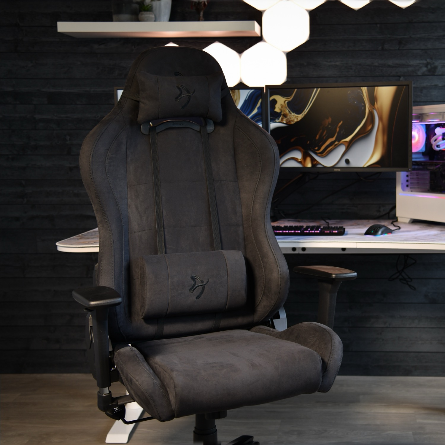 Una sedia da gioco grigio scuro è di fronte a una scrivania con un computer. Lo sfondo comprende un muro nero e luci.