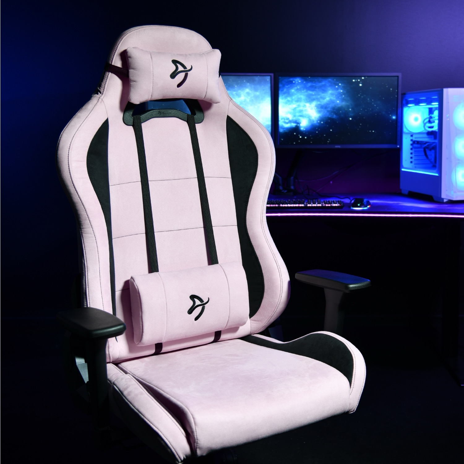 Roze gamingstoel met zwarte accenten. Twee monitoren en een computertoren op de achtergrond.