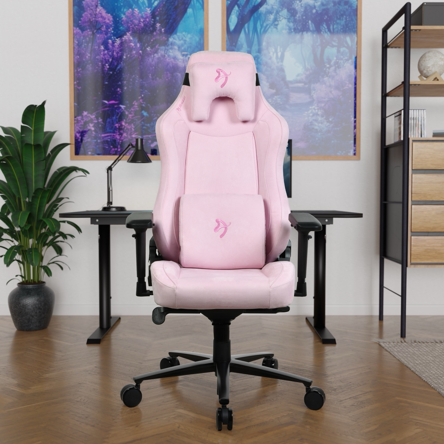 Una sedia da gaming rosa è al centro. Sullo sfondo una scrivania e una mensola, con quadri sul muro. Una pianta a sinistra.
