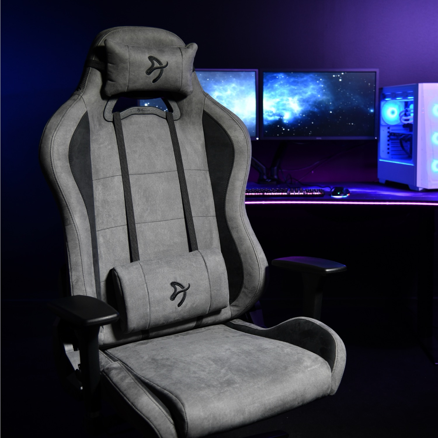 Ein grauer Gaming-Stuhl steht in einem dunklen Raum. Im Hintergrund sind zwei Monitore und ein Computer zu sehen.