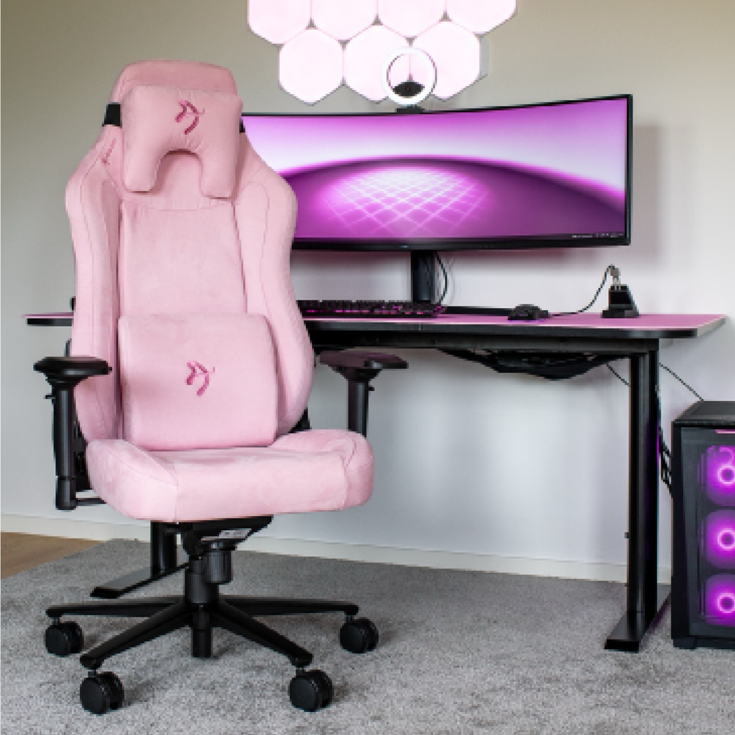 Sedia da gaming rosa, scrivania e configurazione computer. La sedia ha logo sul poggiatesta e supporto lombare. Il monitor mostra un motivo a griglia.