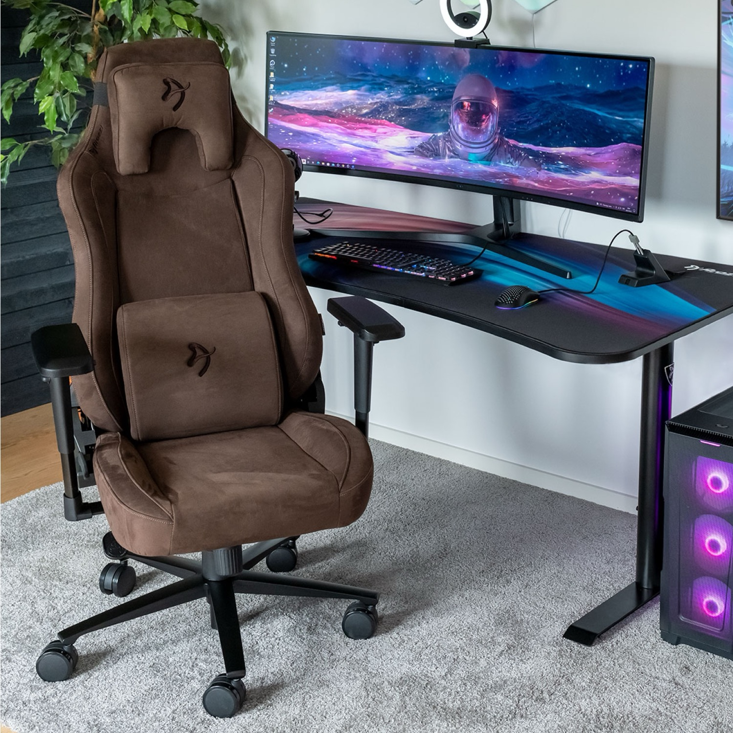Ein brauner Gaming-Stuhl neben einem Schreibtisch mit großem Monitor. Rechts ein Computer-Turm. Der Hintergrund ist eine weiße Wand.