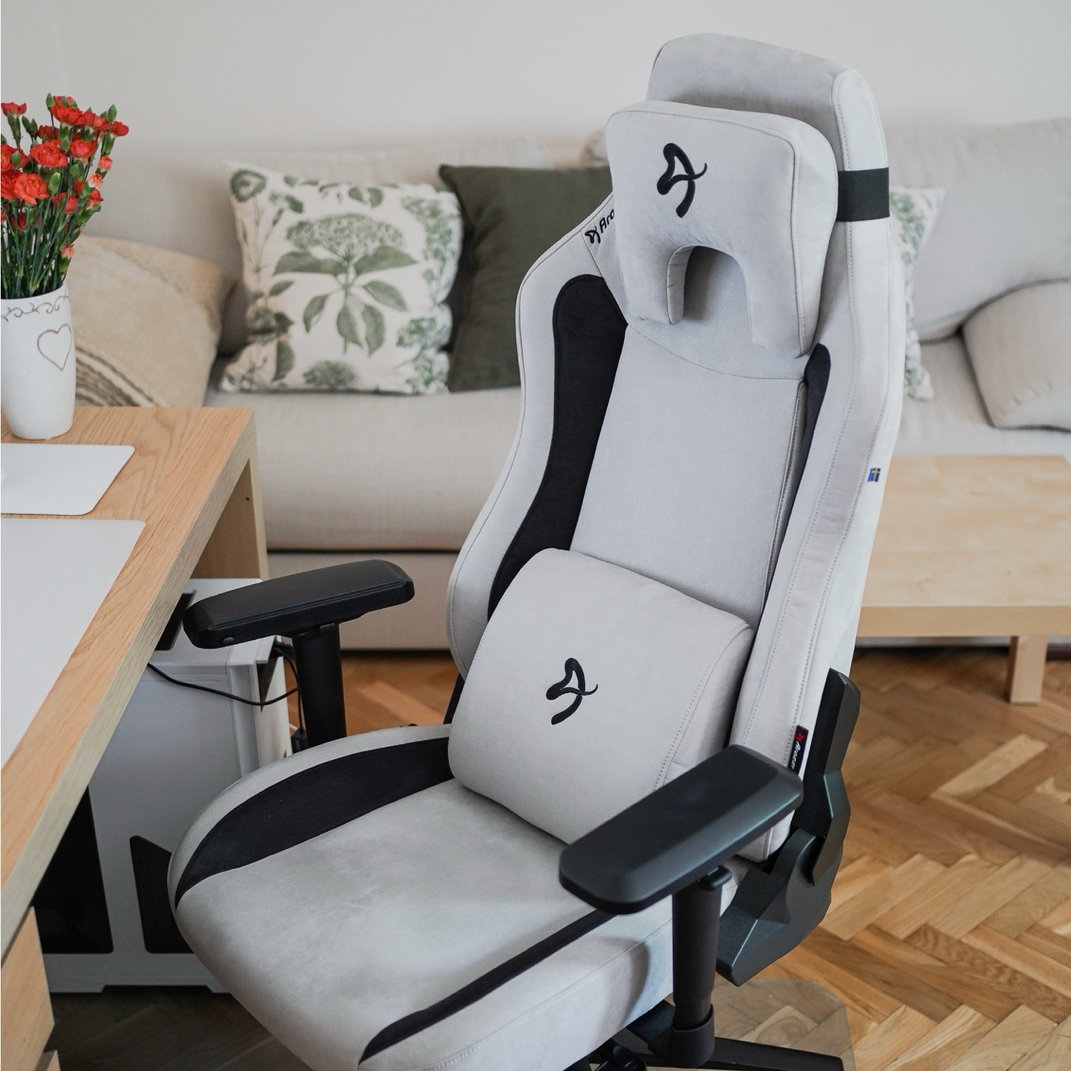 Ein grauer und schwarzer Gaming-Stuhl in einem Raum. Ein Tisch mit Blumen und ein Sofa sind im Hintergrund. Holzboden.