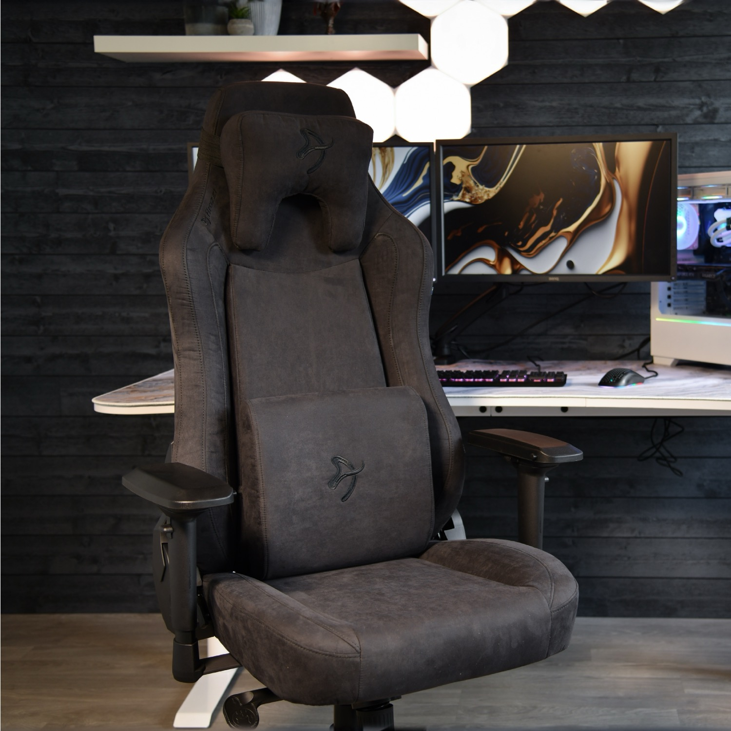 Een donkergrijze gamestoel voor een bureau met een computermonitor. De achtergrond heeft een donkere houten muur en zeshoekige lichten.
