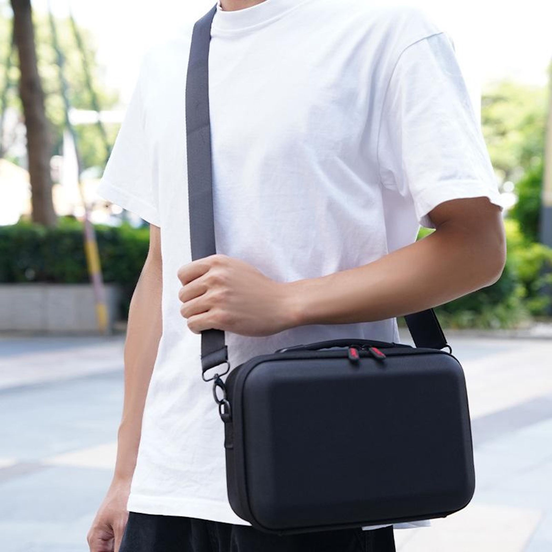 Eine Person trägt ein weißes T-Shirt und hält eine schwarze Tasche mit grauem Riemen. Der Hintergrund ist mit Bäumen verschwommen.
