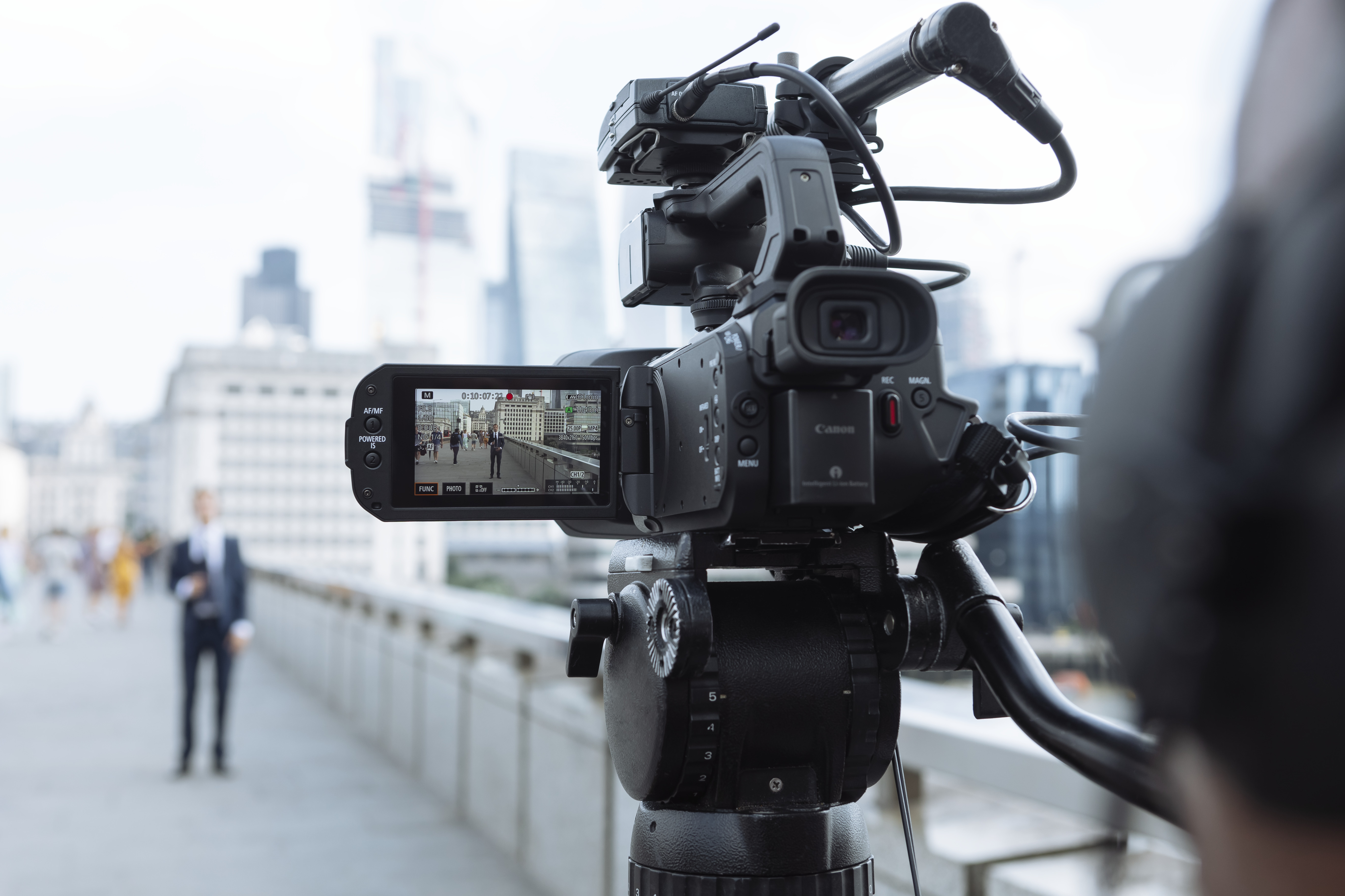 Eine Kamera auf einem Stativ filmt einen Mann im Anzug auf einer Brücke. Gebäude sind im Hintergrund.