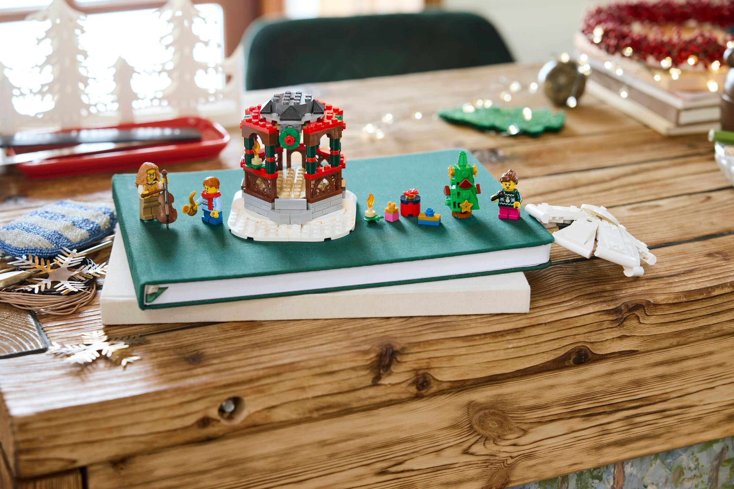Auf einem Holztisch sind Lego-Weihnachtsdekorationen, darunter ein Pavillon, ein Baum und Figuren, auf einem Buch zu sehen.