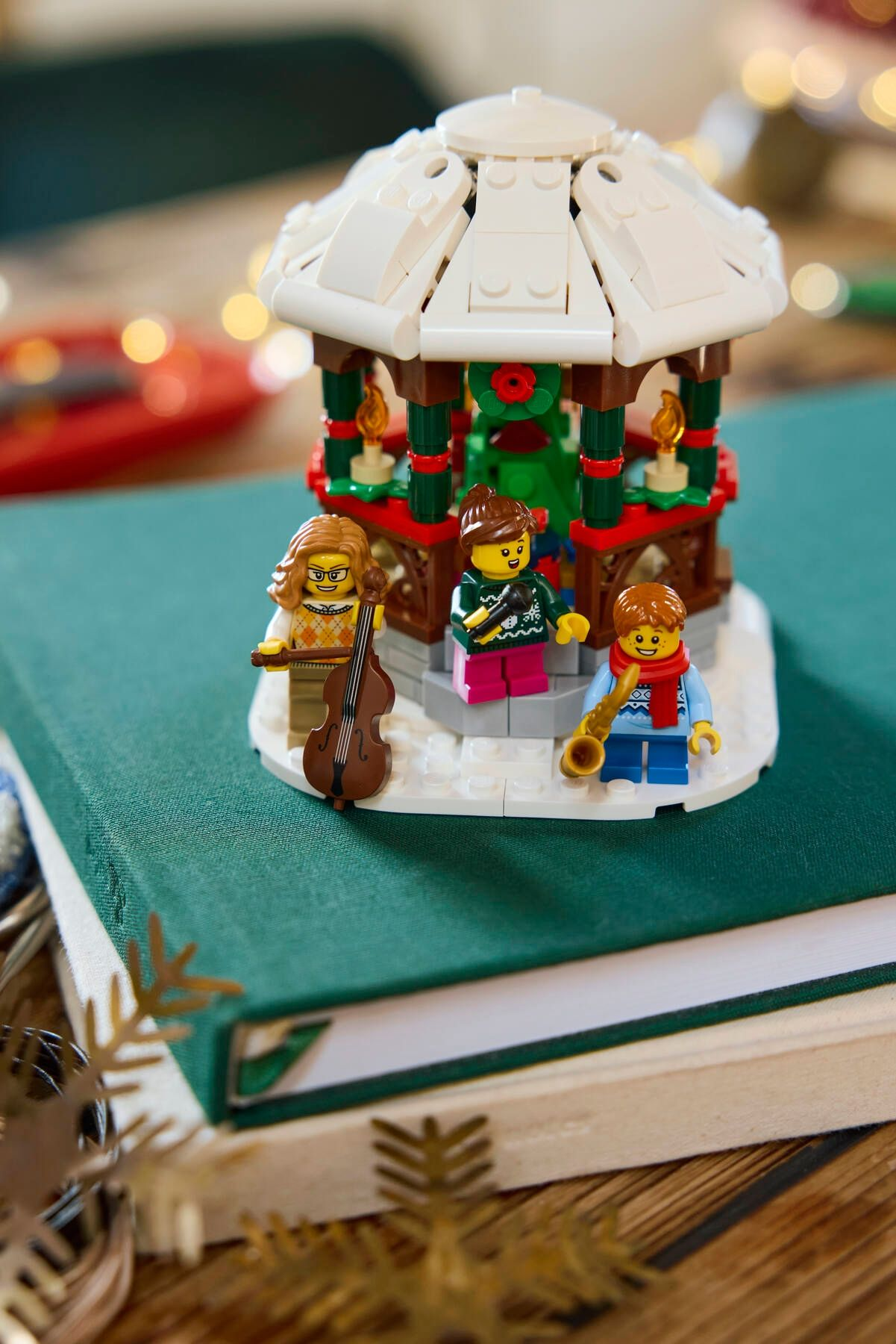 Weihnachtliche Lego-Szene mit Musikern, Karussell und grünem Buch. Figuren spielen Instrumente. Festliche, warme Töne.