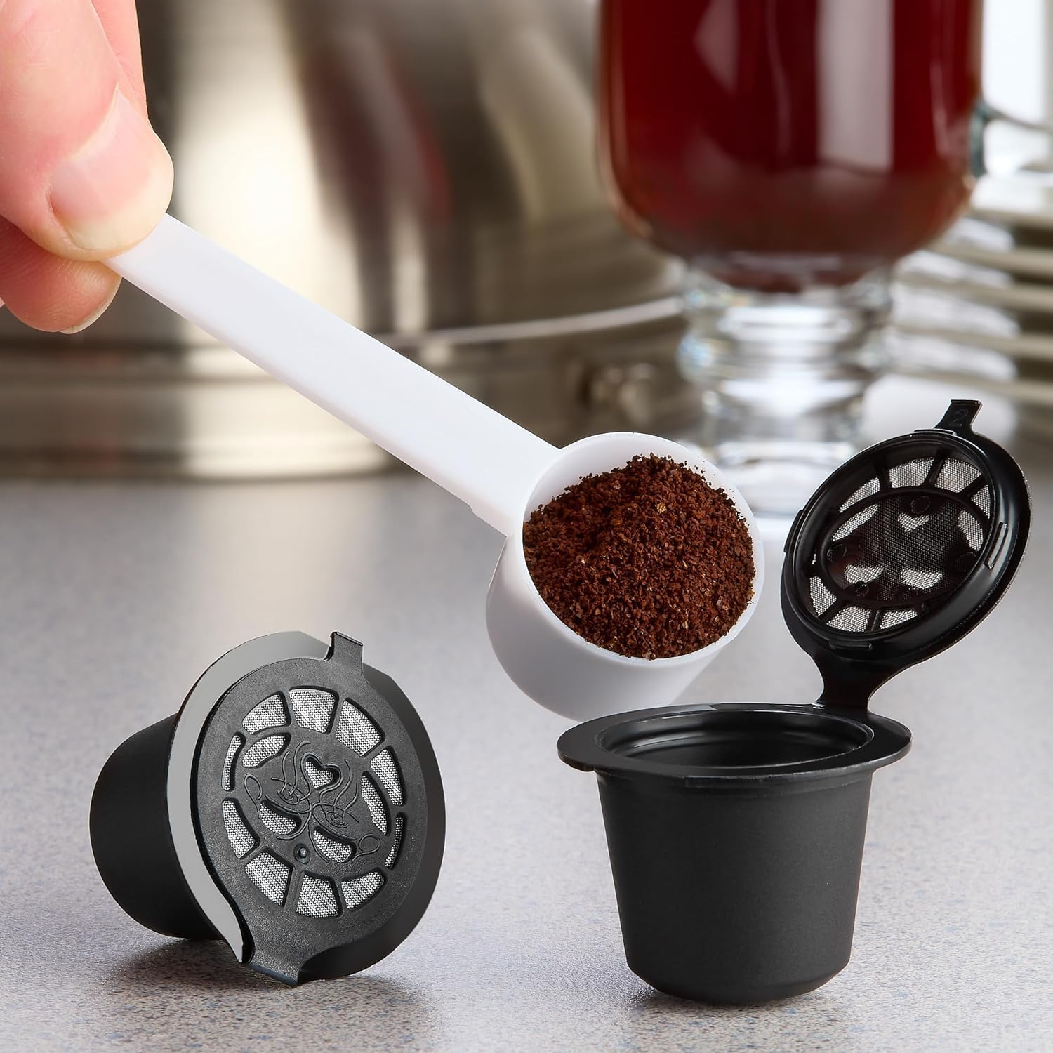 Eine Hand hält einen Löffel mit Kaffeepulver. Im Hintergrund befinden sich zwei schwarze Kaffeepads und ein Glas Kaffee.