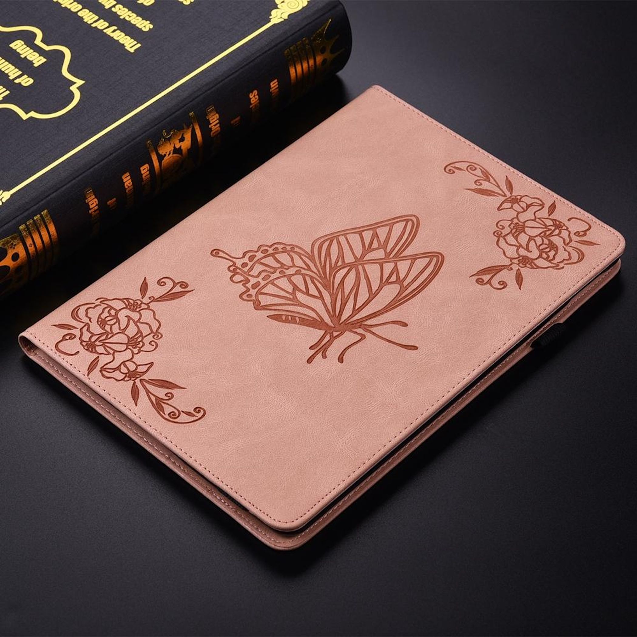 Una funda rosa para tableta con diseño de mariposa y flores. Un libro en el fondo.