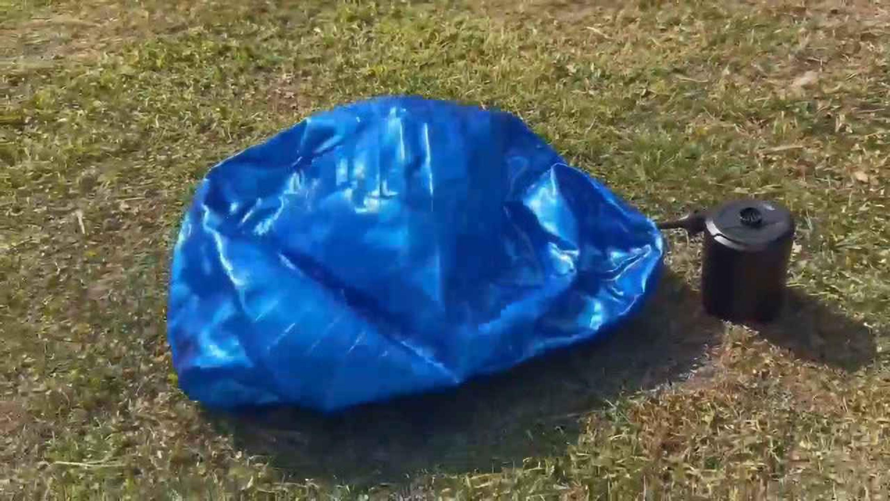 Ein entleertes blaues aufblasbares Objekt liegt auf grünem Gras, neben einer schwarzen Luftpumpe. Das Gras ist leicht trocken.