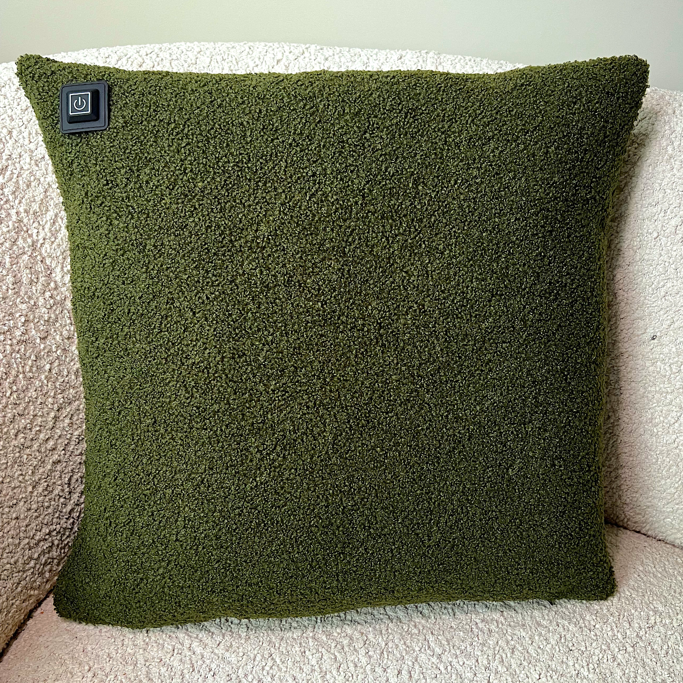 Ein grünes, strukturiertes Kissen mit einem Einschaltknopf liegt auf einer strukturierten, cremefarbenen Oberfläche. Das Kissen ist quadratisch.