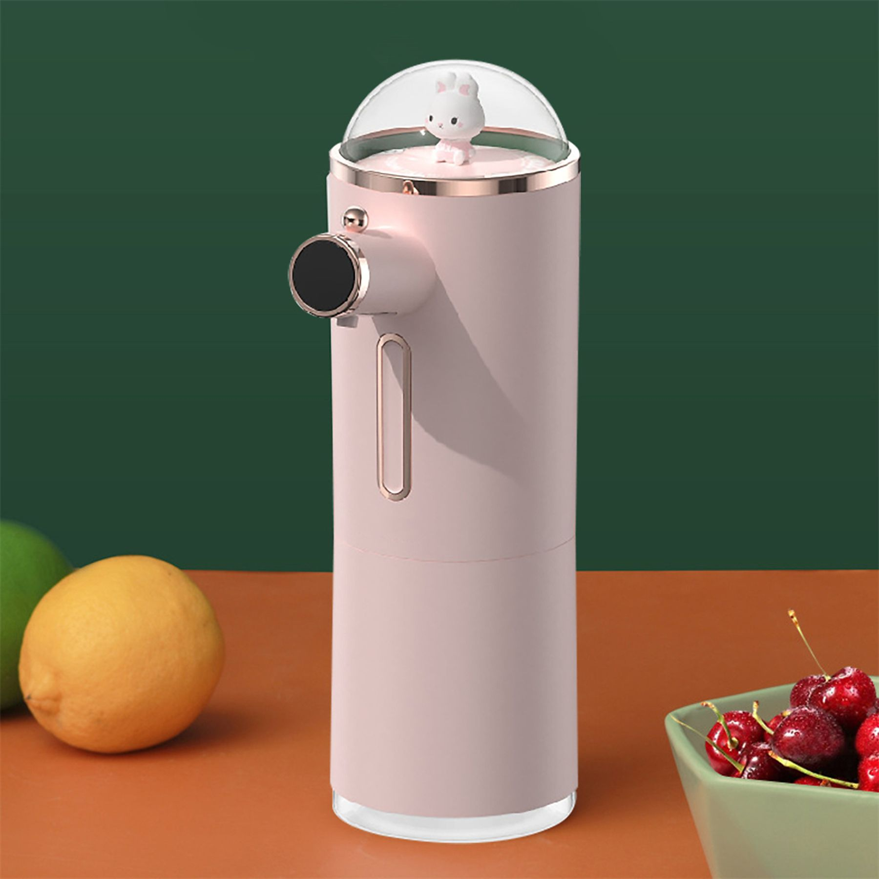 Ein rosa Seifenspender mit einem Häschen oben, Zitrone und Kirschen auf dem Tisch, grüner und orangefarbener Hintergrund.