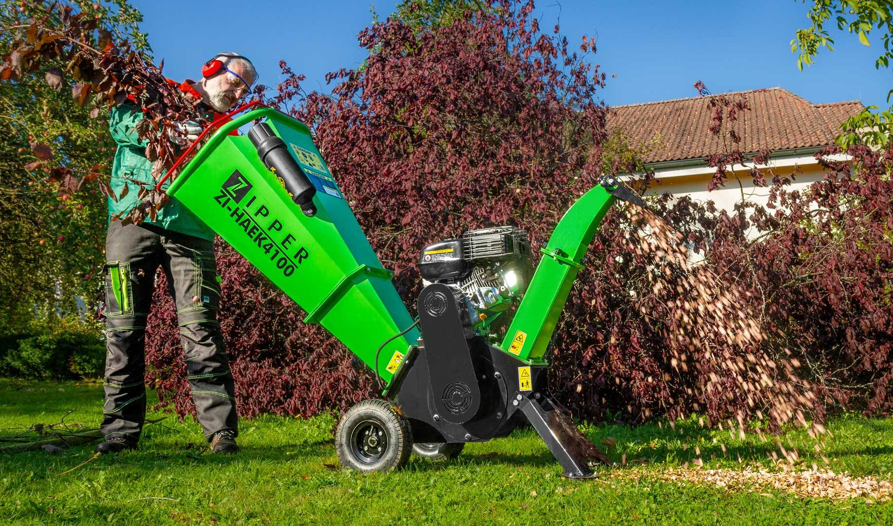 Ein Mann benutzt einen Holzhacker. Grüne Maschine mit Zipper-Logo. Gehacktes Holz spritzt heraus. Hintergrund hat Bäume und ein Haus.