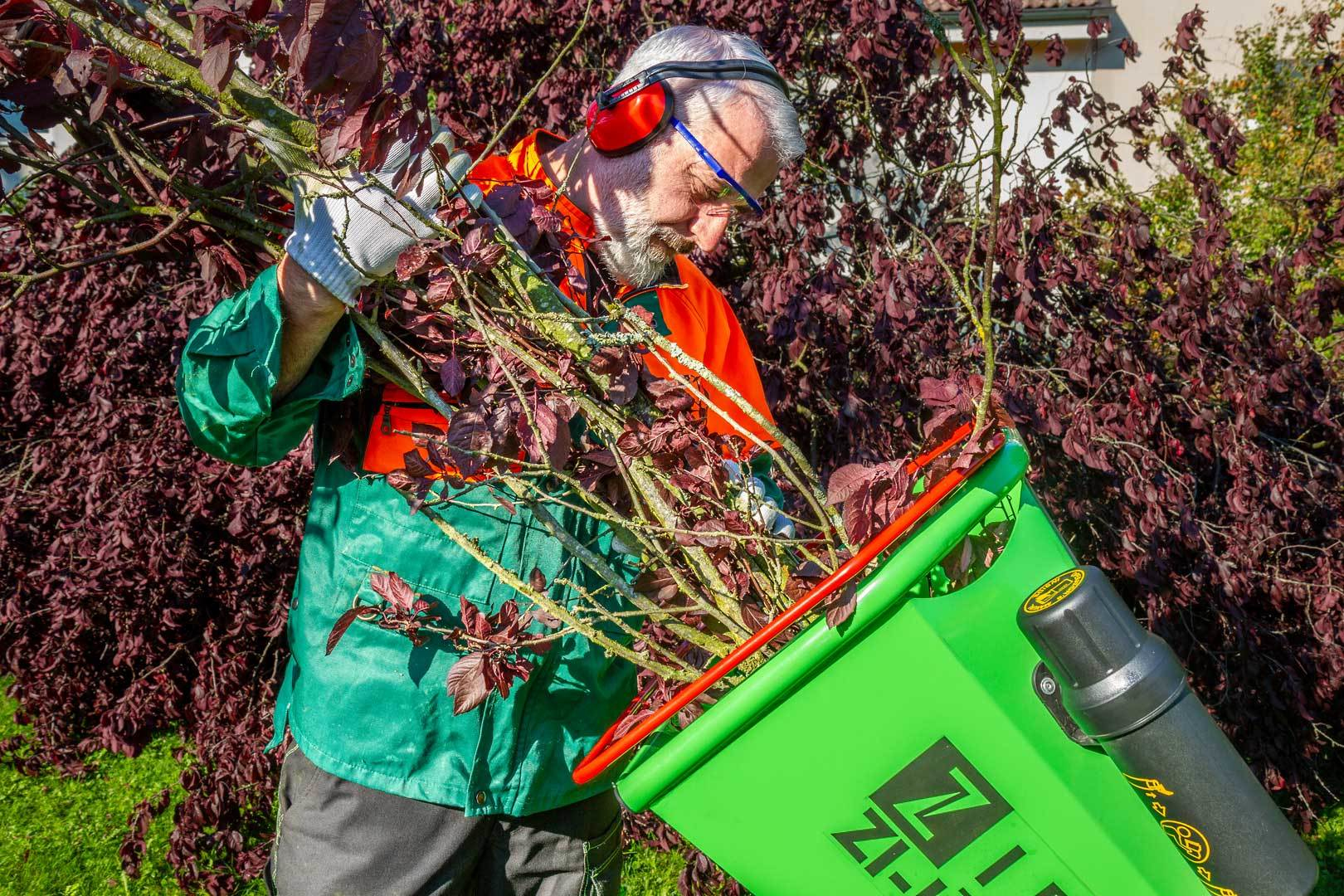 Ein Mann in Schutzkleidung füttert Äste in einen grünen Häcksler. Rote Blätter und Büsche sind im Hintergrund.