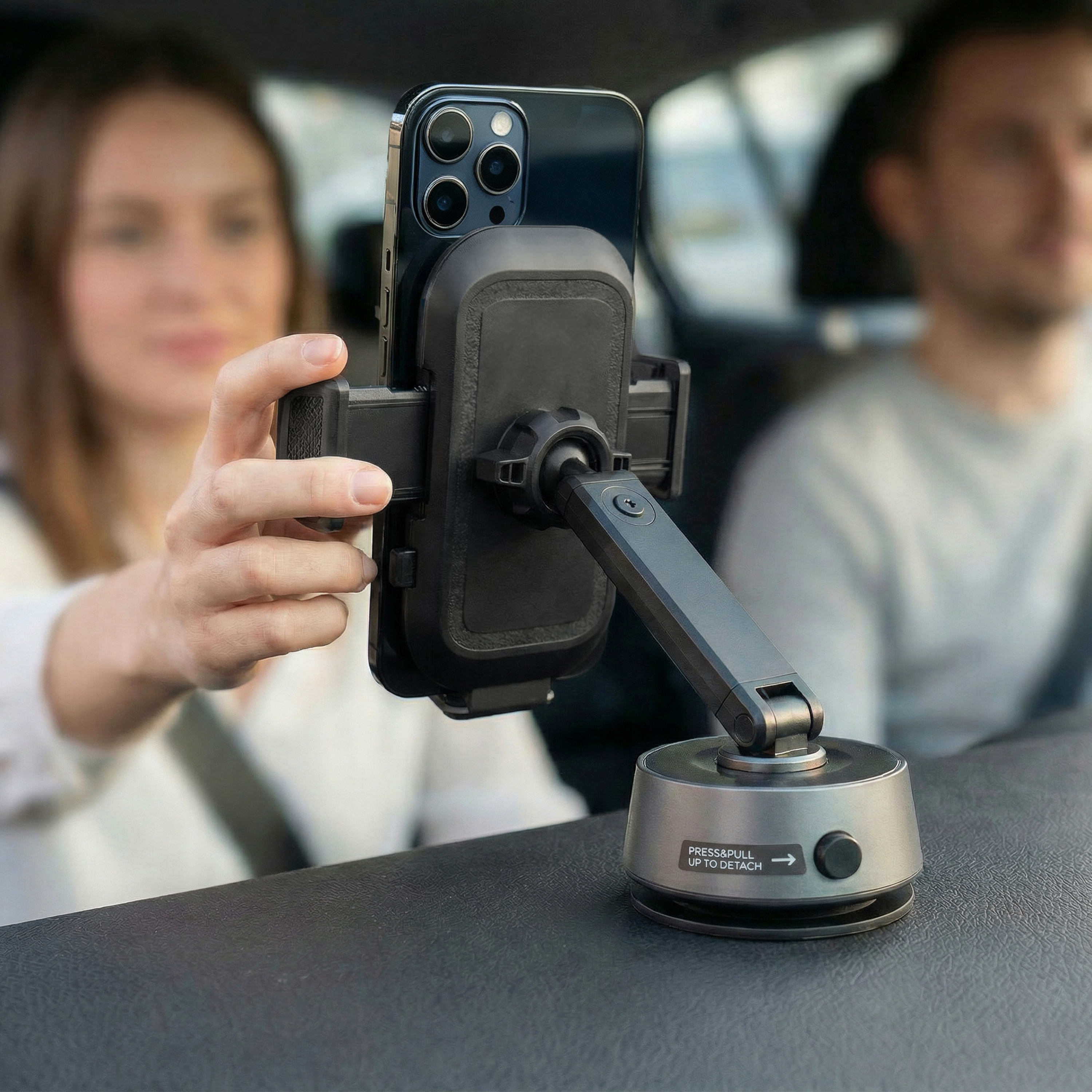 Een telefoon in een autohouder wordt door een vrouw vastgehouden. Een man is wazig op de achtergrond. De houder is zwart en grijs.