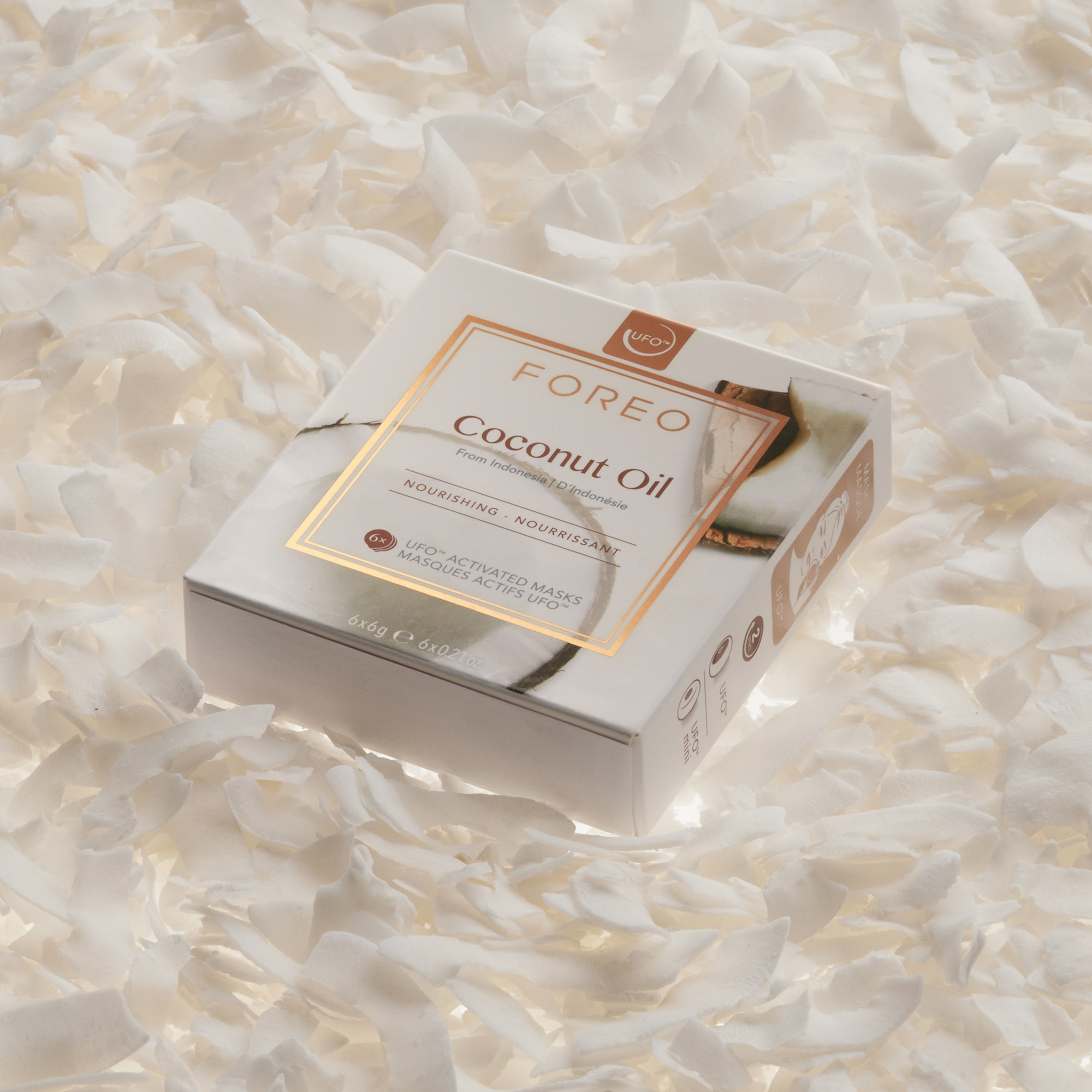 Beyaz hindistan cevizi gevreği üzerinde bir kutu Foreo Coconut Oil maskesi. Kutu, altın renkli metinle beyazdır.
