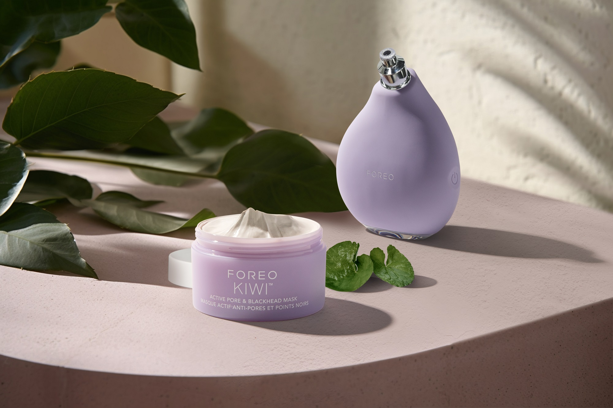 Mor bir Foreo Kiwi cihazı ve maske kavanozu, yapraklarla pembe bir yüzey üzerinde duruyor. Kavanoz beyaz bir maske içeriyor.