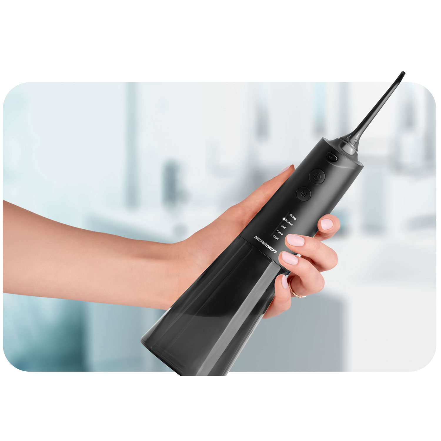 Dłoń trzyma czarny irygator do jamy ustnej. Irygator ma przyciski i dyszę. Tło to rozmyta scena łazienkowa.