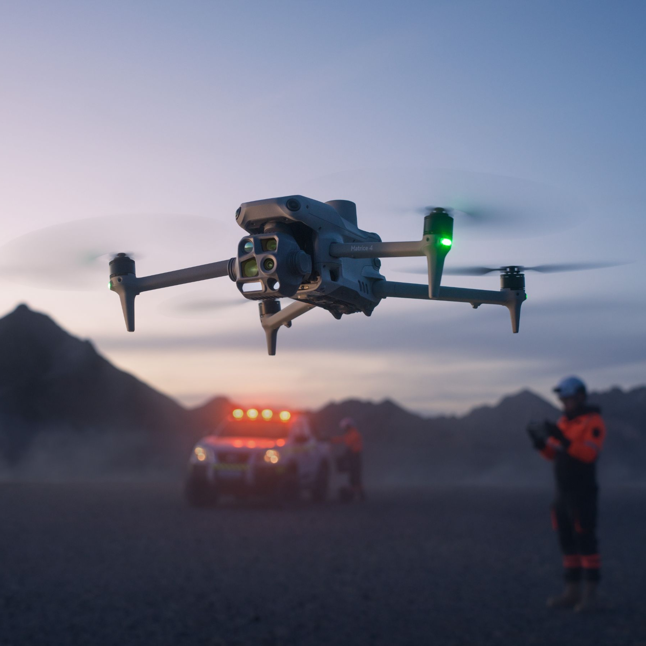 Drone in volo, veicolo di soccorso con luci e una persona sullo sfondo, montagne e cielo.