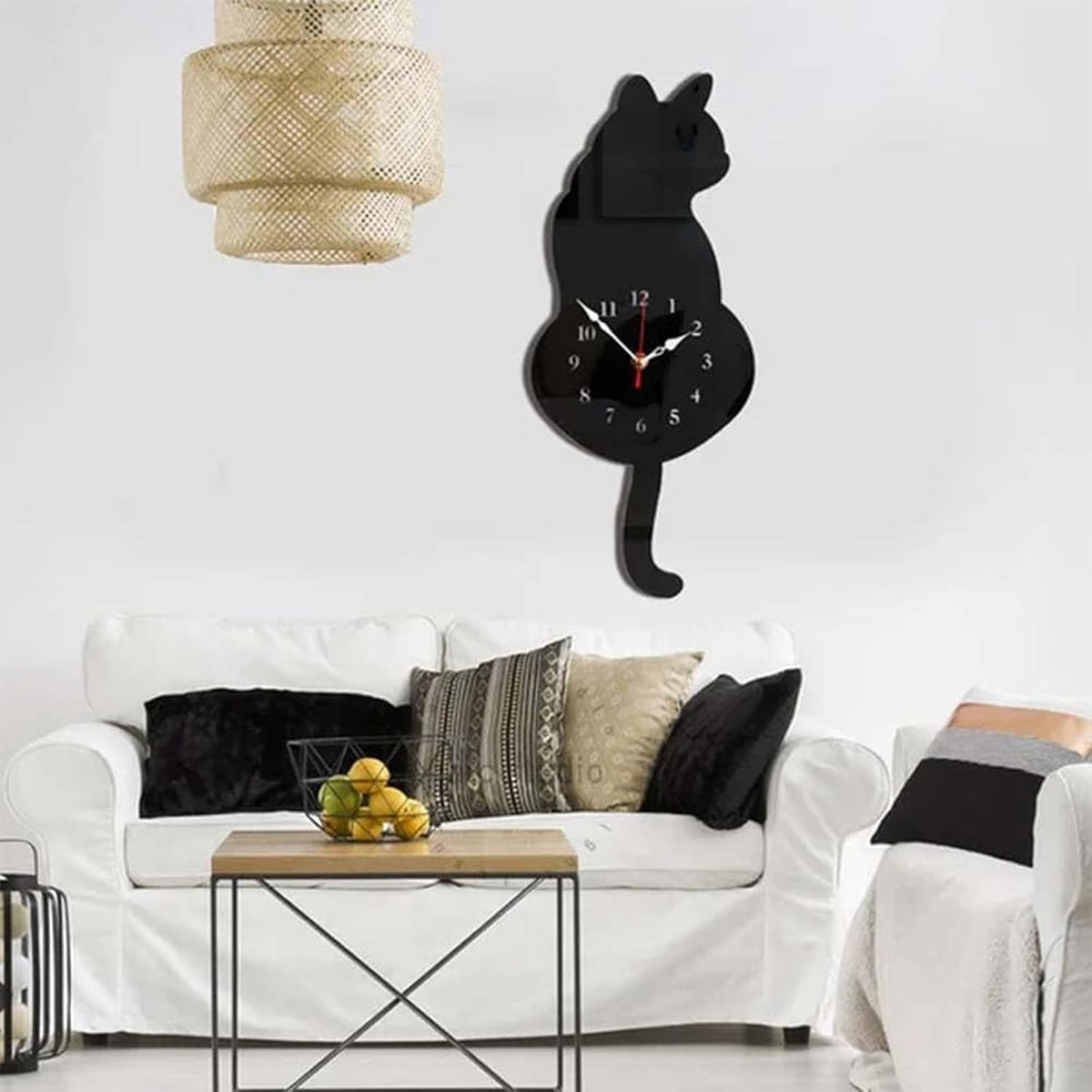 Un orologio a forma di gatto nero su una parete bianca. Sotto, un divano bianco con cuscini, un tavolo di legno e una lampada intrecciata.