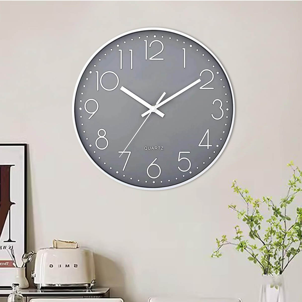 Un orologio al quarzo grigio su un muro, con lancette e numeri bianchi. Un tostapane e una pianta in primo piano.