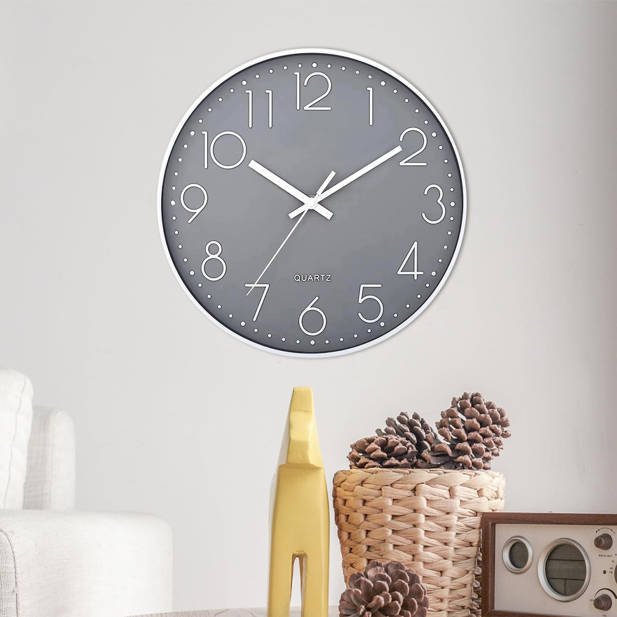 Un orologio da parete grigio con numeri e lancette bianche. Sotto, una statua di cavallo giallo, un cestino di vimini con pigne e una radio.
