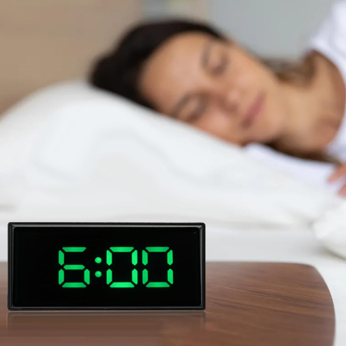 Ein Wecker zeigt 6:00 Uhr in grünen Ziffern an. Im unscharfen Hintergrund schläft eine Frau auf einem weißen Kissen.