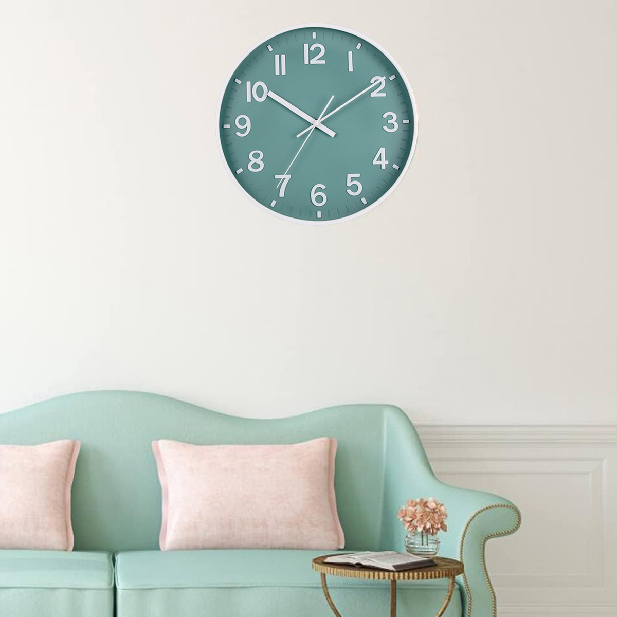Un orologio verde acqua è appeso a una parete bianca sopra un divano verde acqua con cuscini rosa. Un tavolino ha fiori e un libro.
